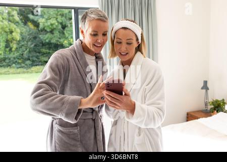 Ältere Mutter und Tochter stehen in einem hellen Schlafzimmer mit weichen Bademänteln und überprüfen das Smartphone Stockfoto