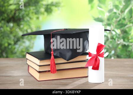 Graduierungskappe, Diplom und Bücher auf Holztisch vor verschwommenem Hintergrund, Großaufnahme Stockfoto