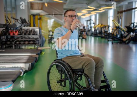 Behindertengerechter Sportler im Rollstuhl, der während des leistungsstarken Workouts im Fitnessstudio ruht und Wasser aus der Flasche trinkt. Stockfoto