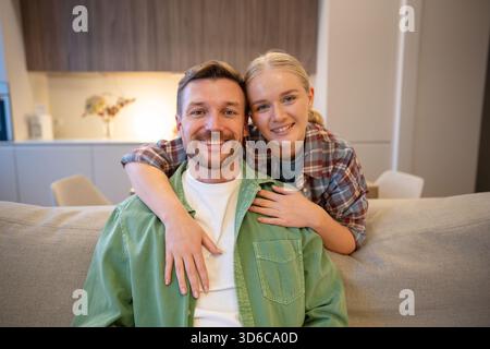 Fröhliches, nettes Paar von Mann und Frau, die sich umarmen und lächeln und sich vor der Kamera im gemütlichen Wohnzimmer posieren. Stockfoto