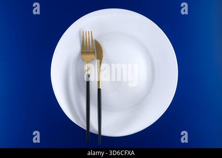 Elegantes, elegantes Geschirr mit weißem Teller und stilvollem goldenem Besteck vor einem leuchtend blauen Hintergrund. Perfekt für kulinarische Zwecke Stockfoto