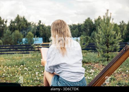 Eine junge kaukasische Frau mit langen, welligen blonden Haaren sitzt draußen und liest ein Buch in einem Dorf umgeben von Grün und Bäumen. Stockfoto
