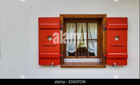 Nahaufnahme der roten Holzfensterläden mit einem herzförmigen Ausschnitt und weichen weißen Vorhängen im Holzrahmen im schweizer Chalet-Stil Stockfoto