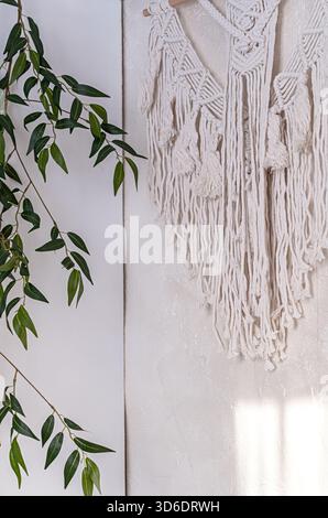Moderne Einrichtung mit handgefertigter Makramee-Wandaufhängung und grünen Ästen. Gemütlicher Boho-Stil, detailgetreues Kunsthandwerk, strukturierte Wand, handwerkliches Dekor. Stockfoto