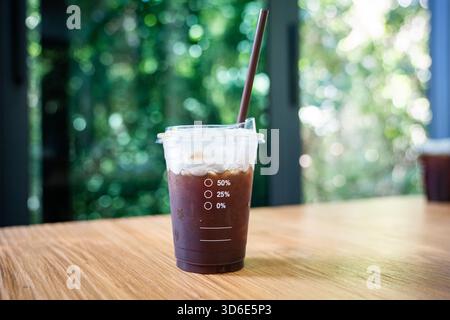 Auf einem Holztisch steht eine Plastiktasse mit eiskaltem schwarzem Kaffee, die klare Messmarkierungen und einen braunen Strohhalm zeigt, der mit Kondenswasser bedeckt ist. Stockfoto