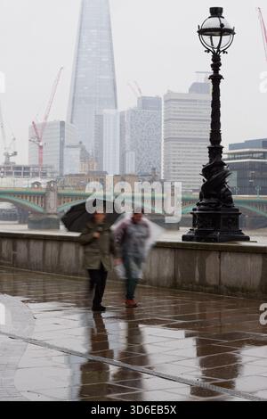 Eine regnerische Hauptstadt von London an einem nassen Tag. Neblig und regnerisch! Zwei Leute laufen an der Themse entlang. Der Splitter in der Ferne Stockfoto