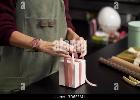 Kaukasische junge Erwachsene Frau bindet rosa Band an Geschenkbox am Tisch, Hände sichtbar, Bogen arrangieren, Präsenzgeschenk im Arbeitsbereich mit Verpackungsmaterialien und dekorativen Gegenständen in der Nähe vorbereiten Stockfoto
