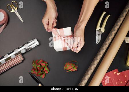 Kaukasische Frau mittleren Alters bindet rosa Band auf Geschenkschachtel, umgeben von Geschenkpapier, Schere, Bändchen und Weihnachtsdekorationen auf dunklem Tisch, um das Geschenk vorzubereiten Stockfoto