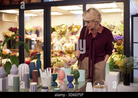 Weiße Frau mittleren Alters arrangiert dekorative Vasen auf dem Tisch im Blumenladen, steht neben der Ausstellung von bunten Blumen und Pflanzen und konzentriert sich auf die Organisation von Blumenarrangements Stockfoto