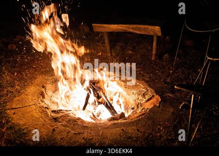 Ein glühendes Lagerfeuer umgeben von Natur, das warmes Licht gegen den Nachthimmel im Wald wirft. Eine rustikale Holzbank und Stühle bilden eine gemütliche Sitzecke Stockfoto