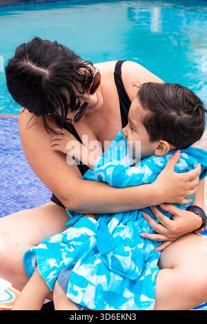 Eine fröhliche Mutter umarmt ihren kleinen Sohn am Pool, er ist in ein leuchtendes blaues Handtuch gewickelt. Die Szene fängt Wärme, Glück und das Wesen von fami ein Stockfoto