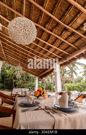 Ein wunderschön gestalteter Essbereich am Strand unter einem traditionellen Palapadach in Cozumel, mit Holzmöbeln, eleganten Tischdekorationen und Tro Stockfoto