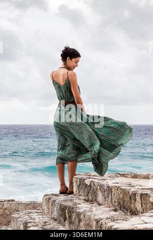Rückansicht einer Frau, die auf Felsen in Cozumel steht und an einem bewölkten Tag ein fließendes grünes Kleid trägt. Stockfoto
