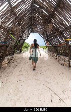 Rückansicht einer nicht erkennbaren Frau mit dunklem Haar, die durch eine traditionelle mexikanische Hütte aus Holz und getrockneten Palmblättern geht, auf t gefangen Stockfoto