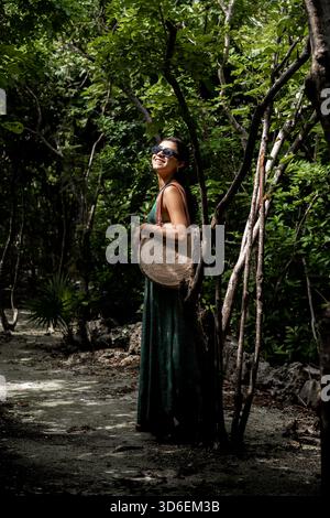 Eine junge Frau in langem Kleid und Sonnenbrille lehnt sich an einen Baum und lächelt im üppigen Grün des tropischen Waldes von Cozumel. Sie trägt eine Tradition Stockfoto