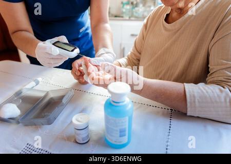 Eine Krankenschwester in Handschuhen testet den Blutzuckerspiegel einer älteren Frau zu Hause mit einem Blutzuckermessgerät, wobei Diabetesmedikation sichtbar ist Stockfoto