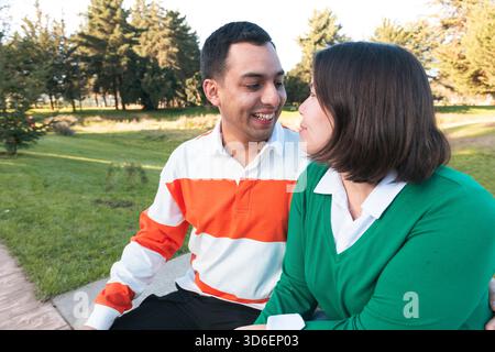 Ein fröhliches, junges lateinisches Paar, das eng zusammen sitzt und sich in einer sonnigen Parklandschaft amüsiert Stockfoto
