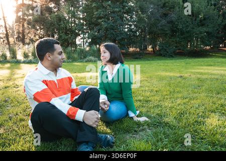 Ein junges Latino-Paar sitzt auf grünem Gras in einem Park und genießt ein angenehmes Gespräch mit warmem Sonnenlicht, das durch die Bäume filtert Stockfoto