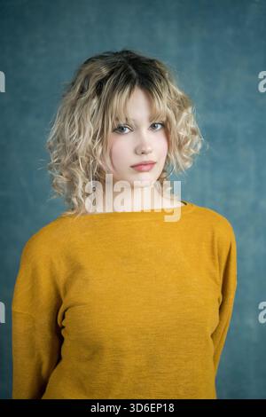 Eine junge Frau mit lockigen blonden Haaren posiert selbstbewusst in einem gelben Pullover vor einem strukturierten blauen Hintergrund, der lässigen Stil und entspannte Elega zeigt Stockfoto