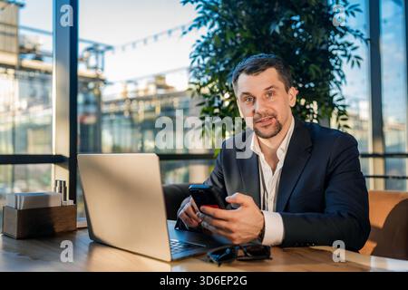 Ein Geschäftsmann im Anzug arbeitet in einem hellen Café an einem Laptop und hält ein Smartphone. Das Tageslicht aus großen Fenstern beleuchtet die moderne, entspannte Einrichtung Stockfoto
