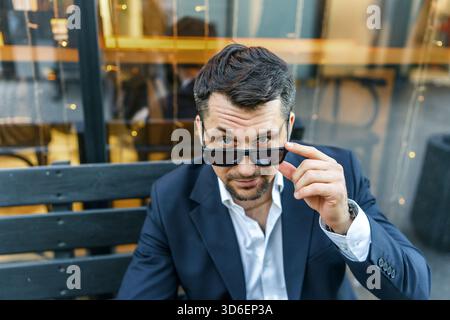 Ein selbstbewusster Geschäftsmann in einem raffinierten Anzug passt seine Sonnenbrille an, während er draußen auf einer Bank sitzt. Er strahlt Professionalität und Stil aus, perfekt für den perfekten Look Stockfoto