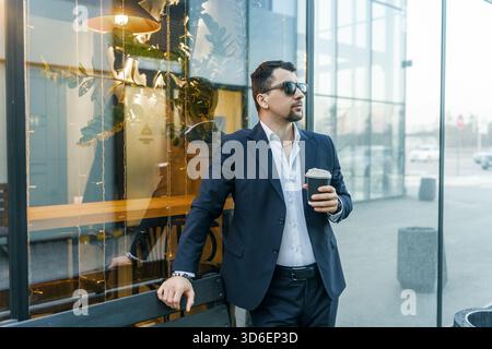 Ein Geschäftsmann in einem eleganten dunklen Anzug und einer Sonnenbrille genießt eine Kaffeepause im Freien und lehnt sich an ein Glasgebäude, das die städtische Umgebung widerspiegelt Stockfoto