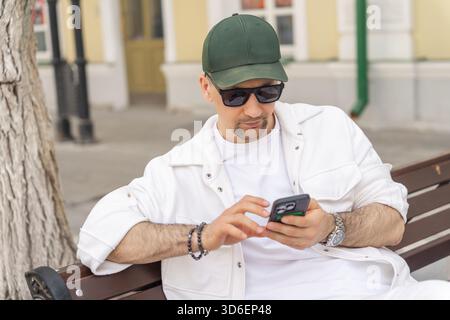 Ein fokussierter Mann in stilvoller weißer Kleidung sitzt auf einer Stadtbank und ist vertieft, auf seinem Smartphone zu surfen. Stockfoto