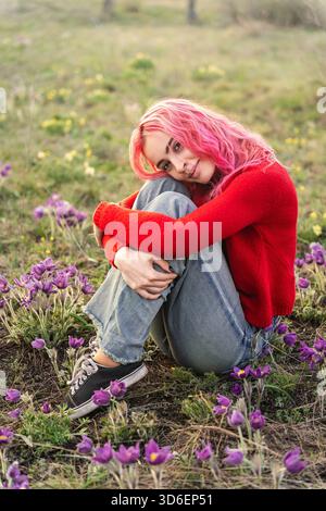 Eine junge Frau mit leuchtend rosa Haaren sitzt friedlich inmitten eines lila Blumenfeldes, trägt einen roten Pullover und Jeans und hält einen ruhigen Moment in n fest Stockfoto