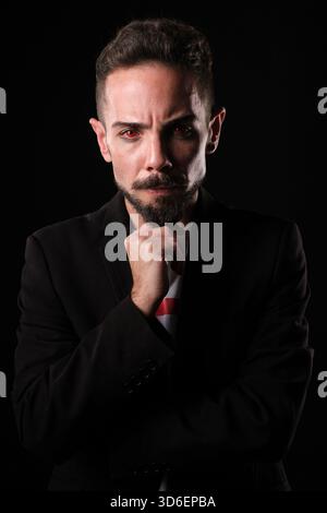 Ein ernsthaft aussehender Mann in einem Anzug mit roten Kontaktlinsen, der vor dunklem Hintergrund steht und sein Kinn auf seiner Hand liegt. Stockfoto