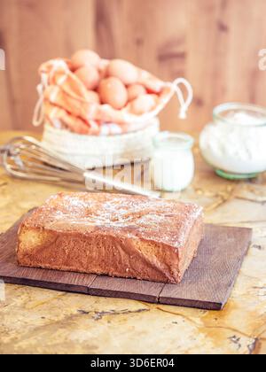 Ein hausgemachter Biskuitkuchen steht auf einem rustikalen Holztisch, der mit Puderzucker bestäubt ist, mit Zutaten wie Eiern, Mehl und Milch in der Nähe, perfekt für baki Stockfoto