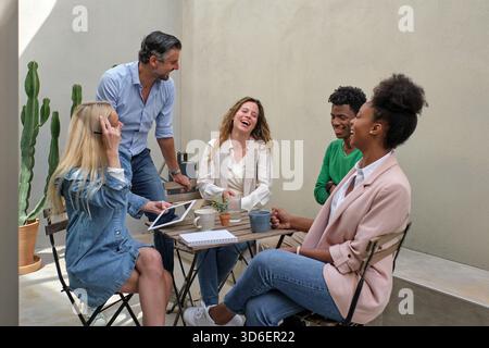 Positive, multirassische Kollegen treffen sich auf der Terrasse um den Tisch und lachen während der Arbeit zusammen Stockfoto