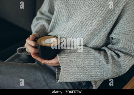 Eine gemütliche Szene, in der eine Person in lässiger Kleidung eine Tasse Kaffee mit komplizierter Latte Art hält. Das entspannte Ambiente strahlt Wärme und Komfort aus Stockfoto