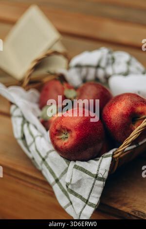 Frische, saftig rote Äpfel in einem rustikalen Webkorb auf einem Holztisch angeordnet, teilweise mit einem grün-weiß karierten Tuch bedeckt. Stockfoto