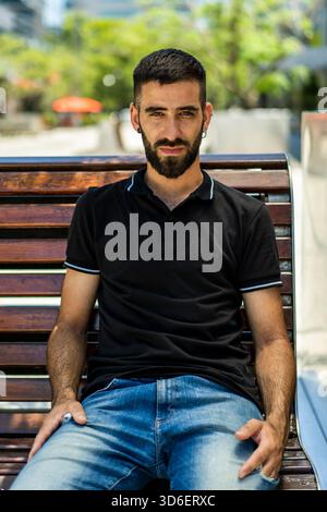 Porträt eines jungen Mannes mit Bart, der bequem auf einer Parkbank sitzt und Ruhe in einer sonnigen städtischen Umgebung ausstrahlt Er trägt ein dunkles Poloshirt PA Stockfoto