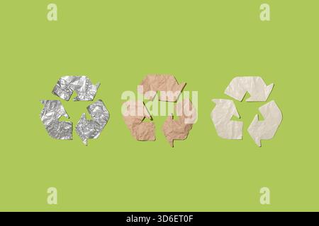 Drei Recyclingsymbole aus Aluminiumfolie, braunem Papier und Kunststoff vor einem grünen Hintergrund, die nachhaltige Praktiken und Öko-fri symbolisieren Stockfoto