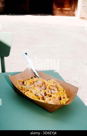 Eine Portion Spaghetti alla Carbonara in einer braunen Pappschale mit einer Metallgabel, die draußen im Sonnenlicht auf einem grünen Tisch steht Stockfoto