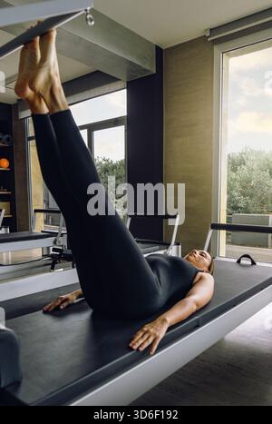 Eine Frau in schwarzer Sportkleidung führt in einem hellen, modernen Studio eine Pilates-Reformer-Beinübung durch. Sie ist fokussiert und fit und fesselt ihren Körper Stockfoto