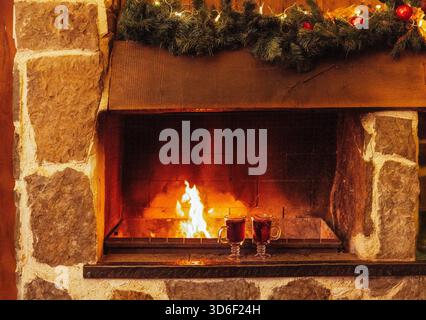 Zwei Gläser mit Glühwein sind neben dem Kamin. Weihnachtsdekoration und Tassen mit einem scharfen Heißgetränk. Offene Feuer- und Steinmauern. Silvester Erholung, Entspannung und Urlaub Stockfoto