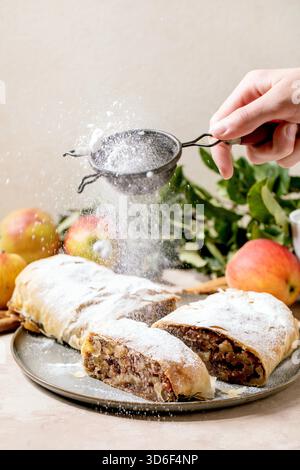 Hausgemachter klassischer Apfelstrudel mit Puderzucker aus dem Sieb in der Hand, auf Keramikplatte mit frischen Äpfeln, grünen Blättern und Zimtstangen darüber. Stockfoto