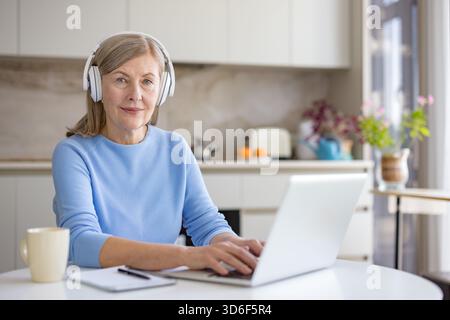 Ältere Frau, die Kopfhörer trägt, während sie auf einem Laptop schreibt, aus der Küche arbeitet oder lernt und moderne Technologien für die Ausbildung oder berufliche Entwicklung nutzt Stockfoto