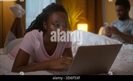 Afroamerikaner Familie verbringen Abend Internet Frau benutzen Laptop Ehemann Handy ethnisch verheiratetes glückliches Paar Bett zu Hause Schlafzimmer Nacht online Stockfoto