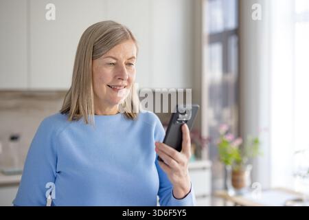 Eine ältere Frau lächelt, während sie ein Smartphone hält und für Videoanrufe verwendet, mit Familie oder Freunden kommuniziert und über Technologie in Verbindung bleibt Stockfoto