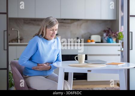 Ältere Frau, die an starken Magenschmerzen und -Beschwerden leidet und ihre Hände über den Bauch hält, während sie an einem Küchentisch sitzt und eine Episode von Verdauungsstörungen erlebt Stockfoto