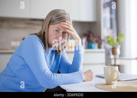 Ältere grauhaarige Frau, die an einem Küchentisch saß und mit der Hand auf der Stirn nach unten schaute, nachdenklich und beunruhigt erschien, unter Kopfschmerzen, Sorgen und Einsamkeit litt Stockfoto