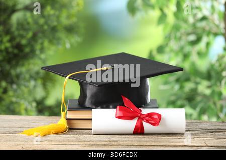 Graduierungskappe, Diplom und Bücher auf Holztisch vor verschwommenem Hintergrund, Großaufnahme Stockfoto