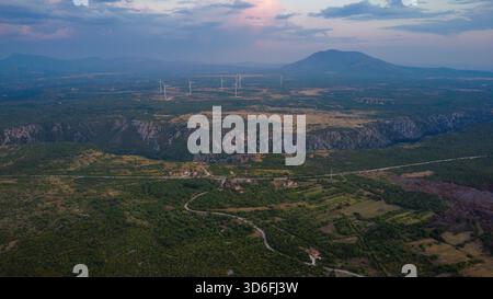 Riesige europäische Landschaft mit modernen Windturbinen und einer steilen Flussschlucht, mit sanftem Gelände und fernen Bergen unter einem dramatischen Abenddämmerhimmel Stockfoto