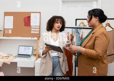Verschiedene weibliche Kollegen, die Stoffmuster und digitale Skizzen auf einem Tablet im Designstudio überprüfen Stockfoto