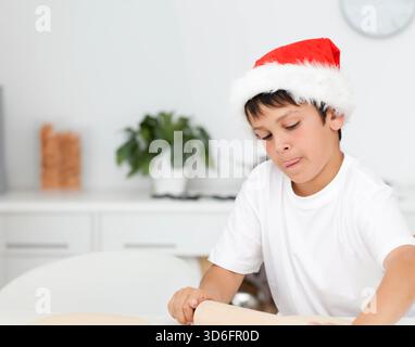Junge Kind, das an einem runden Tisch in der Küche sitzt, trägt Weihnachtsmann-Hut, der Teig mit Holznadel rollt. Festlich, Gebäck, gemütlich, Korb, Kind, hell, Lifestyle Stockfoto