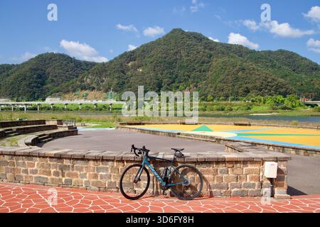 Ein Rennrad lehnt sich an eine niedrige Steinmauer an einem farbenfrohen plaza am Flussufer in Gangchon, mit Hügeln und einem langen Viadukt, das über das Tal sichtbar ist. Stockfoto