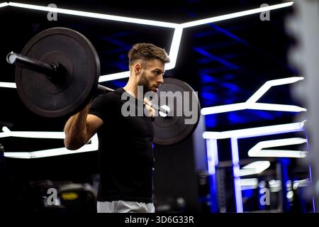Ein Mann führt eine Langhantelhocke in einem stilvollen Fitnessstudio aus, wobei er sich auf Krafttraining konzentriert. Der Fitnessraum verfügt über Neonlichter und ein modernes Design, kreativ Stockfoto